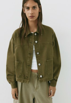 Mladá žena s dlhými hnedými vlasmi, oblečená v overzise olivovo zelenej košeľovej bunde a k nej ladúce nohavice s vysokým pásom cez biely crop top.