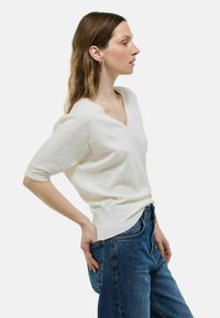 Pull blanc à manches courtes avec un col en V, présentant une texture douce et des accents côtelés subtils, associé à un jean en denim bleu.