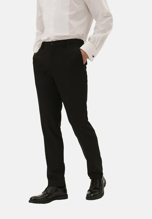 Mężczyzna noszący czarne eleganckie spodnie, czarne eleganckie buty i białą koszulę z mankietami, stoi z rękami w kieszeniach.