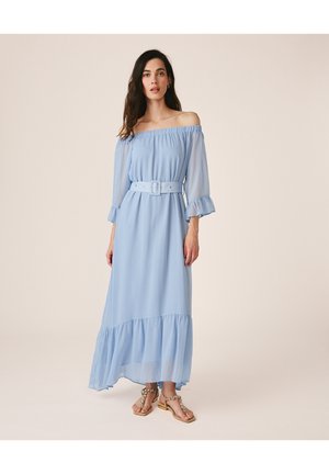 Frau trägt ein hellblaues schulterfreies Maxikleid mit passendem Gürtel und Rüschen am Saum, kombiniert mit flachen Sandalen mit Perlenbesatz.