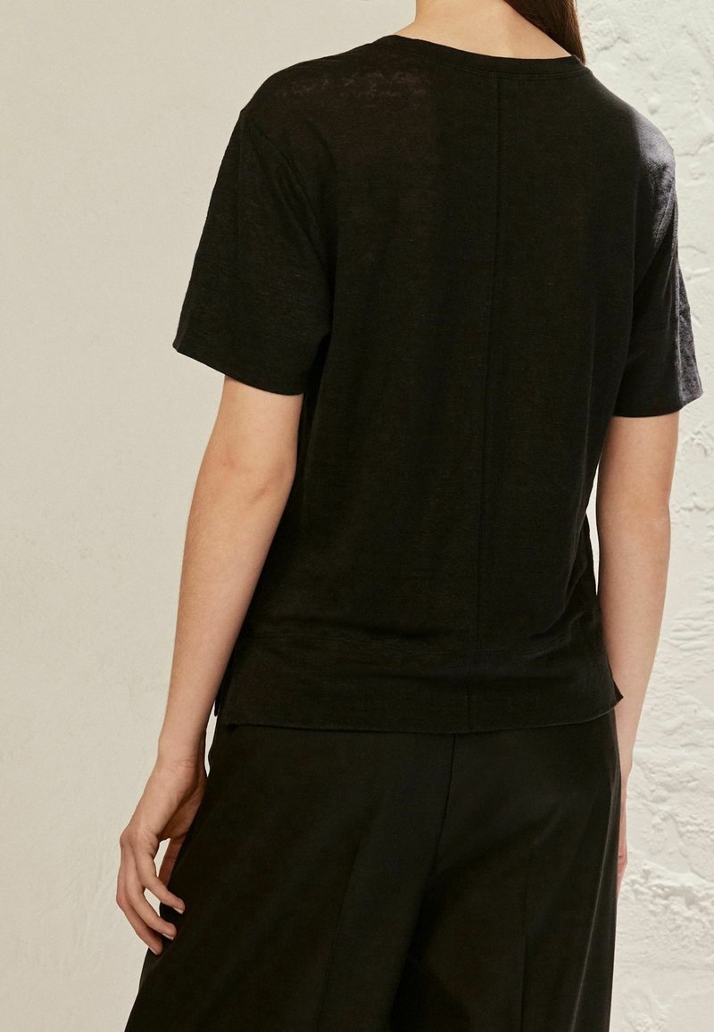 Personne portant un t-shirt noir uni à manches courtes et un pantalon noir, vue de dos sur un fond de mur clair.