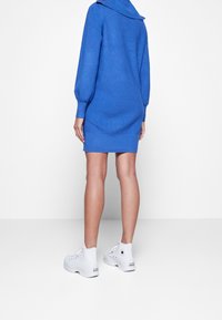 Blå stickad klänning med krage, långa puffärmar och figurnära passform, kombinerad med vita högskor. Slät textur med ribbade detaljer.