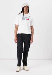 White t-shirt with blue and red "New Balance Athletic Shoes" logo, paired with black trousers and beige athletic shoes. Minimalist design.