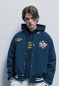 Námořnická modrá varsity bunda s bílými proužkovanými manžetami a límcem, s vyšitými texty a logy v žluté a červené barvě. Nositá přes černou mikinu s kapucí.