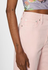 Calças de ganga rosa de cintura alta com bolsos frontais, apresentando acessórios prateados. O tecido tem uma textura suave. Mão com uma tatuagem visível.