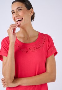 Mujer sonriente con cabello oscuro recogido en un moño, lleva una blusa roja con patrones florales recortados, con la mano cerca de la boca y el brazo cruzado sobre un fondo blanco.