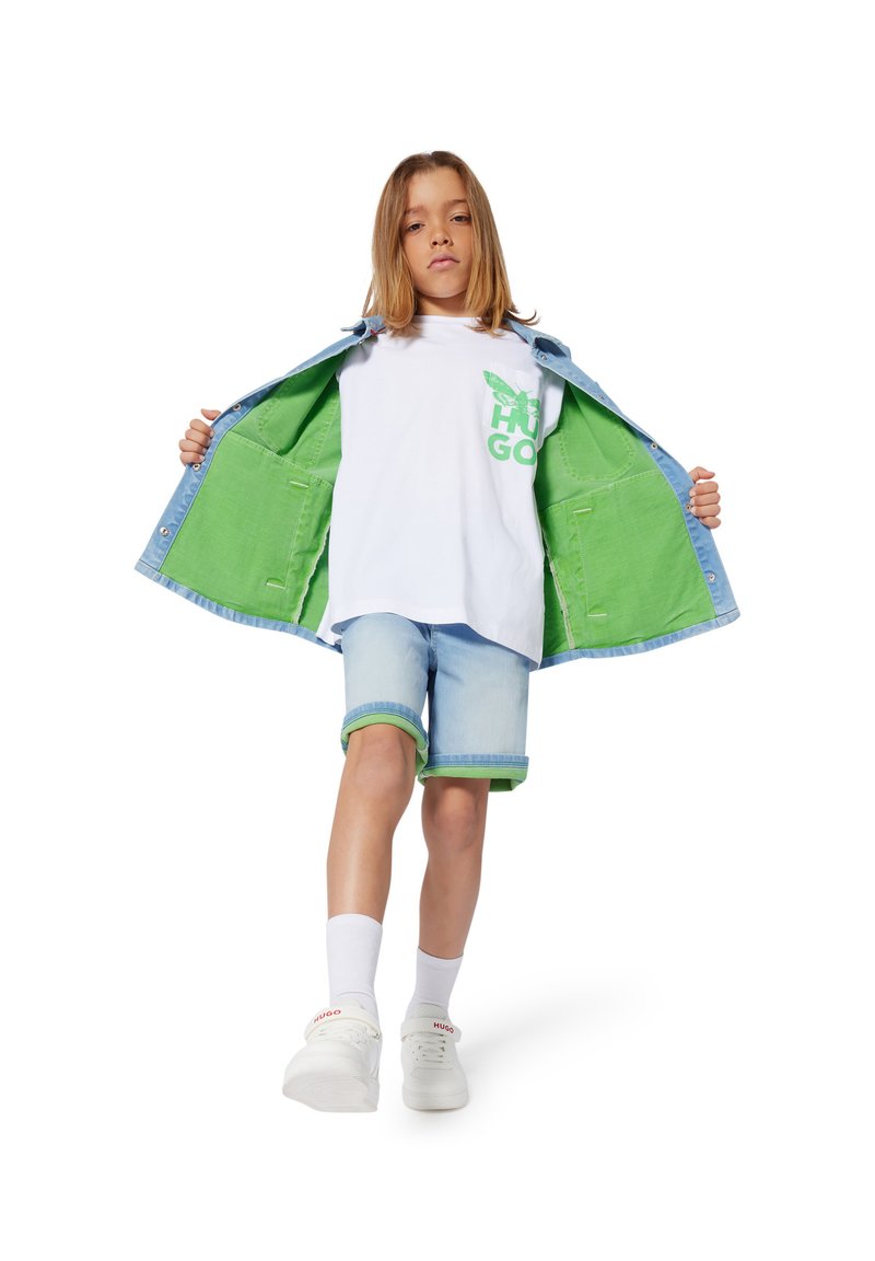 Denim jack met lichtblauwe buitenkant en groene binnenkant, witte T-shirt met groene graphics, lichtblauwe shorts met groene accenten, witte sneakers.