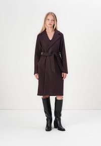 Abrigo largo de lana de color púrpura oscuro con cintura ceñida, cuello de solapa y bolsillos laterales, combinado con botas de cuero negras hasta la rodilla.