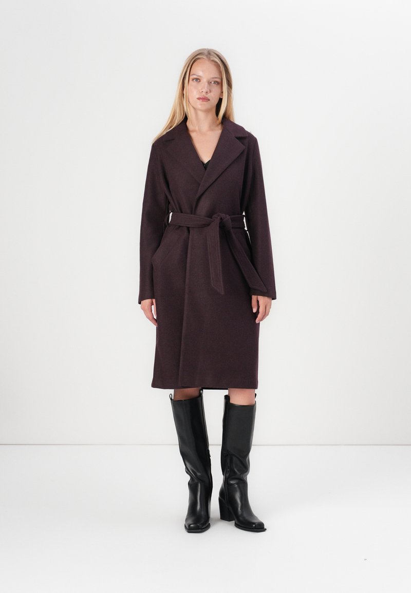 Abrigo largo de lana de color púrpura oscuro con cintura ceñida, cuello de solapa y bolsillos laterales, combinado con botas de cuero negras hasta la rodilla.