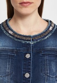 Donna che indossa una giacca di jeans blu scuro con borchie decorative argentate attorno al collo e bottoni visibili sul davanti.