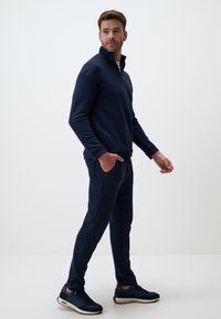 Sweatshirt zippé bleu marine et joggings assortis en tissu doux avec poches latérales et coupe décontractée, accompagnés de baskets noires et blanches.