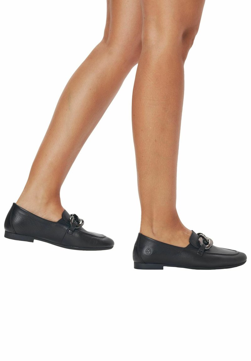 Mocasines de cuero negro con suela plana, que presentan una correa decorativa con un acento metálico circular y un acabado de textura suave.