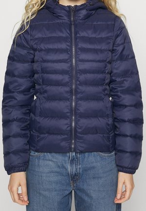 Personne portant une veste matelassée à capuche bleu marine zippée, associée à un jean bleu en denim.