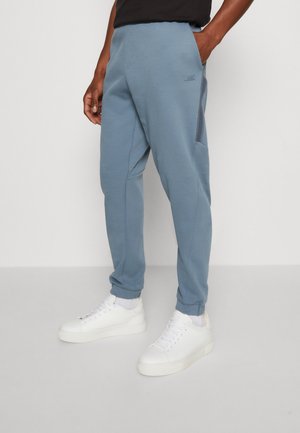 Ein Mann trägt hellblaue, schmal zulaufende Jogginghosen mit seitlichem Mesh-Einsatz und weiße Turnschuhe, steht mit einer Hand in der Tasche.