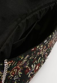 Bolso con estampado floral y forro negro, con una correa de cadena plateada y cierre de botón a presión. Textura de tela visible.