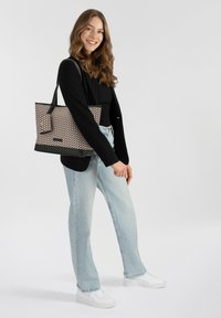 Borsa tote con un motivo geometrico in beige e marrone, dotata di manici neri e fondo piatto. Il modello indossa jeans azzurri chiari e una maglietta nera.