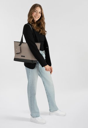 Borsa tote con un motivo geometrico in beige e marrone, dotata di manici neri e fondo piatto. Il modello indossa jeans azzurri chiari e una maglietta nera.