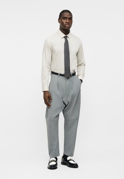 Άνδρας που φοράει ριγωτό κρεμ πουκάμισο, σκούρα γραβάτα, γκρι πλισέ παντελόνια, μαύρα και λευκά μοκασίνια, στέκεται με το ένα χέρι στην τσέπη σε λευκό φόντο.