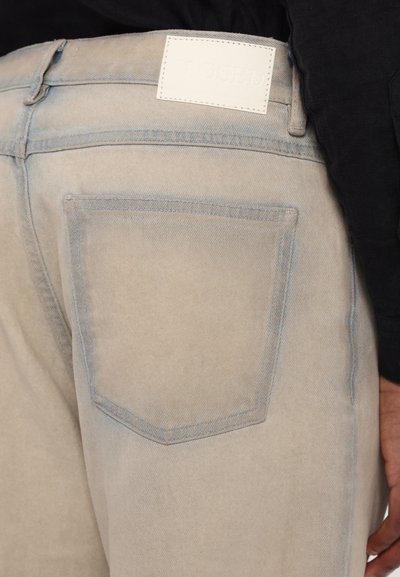Pantaloni in denim beige chiaro con una texture liscia, dotati di una tasca posteriore, cuciture blu a contrasto e un'etichetta del marchio in pelle bianca.