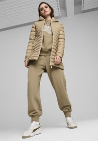 Beige pufferjakke med hætte, med horisontale syninger, parret med en matchende beige hættetrøje og joggers. Hvide sneakers med olivengrønne accenter.