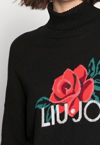 Maglione a collo alto nero con una grafica di una rosa rossa e foglie verdi e un testo stilizzato bianco parzialmente visibile sul petto.