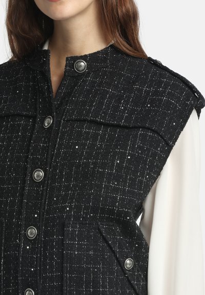 Veste noire texturée avec des coutures métalliques, dotée de gros boutons argentés, d'un col montant et de deux poches avant, portée par-dessus une chemise blanche.