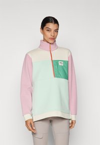 Farverig pullover sweatshirt lavet af blødt stof. Har en høj krave, halvdels lynlås og en brystlomme i grøn med et logo. Farverne inkluderer pink, mintgrøn og creme.