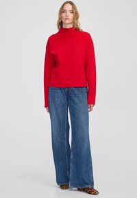 Red ribbed turtleneck sweater with dropped shoulders, paired with blue wide-leg denim jeans and animal print shoes. Plain background.
