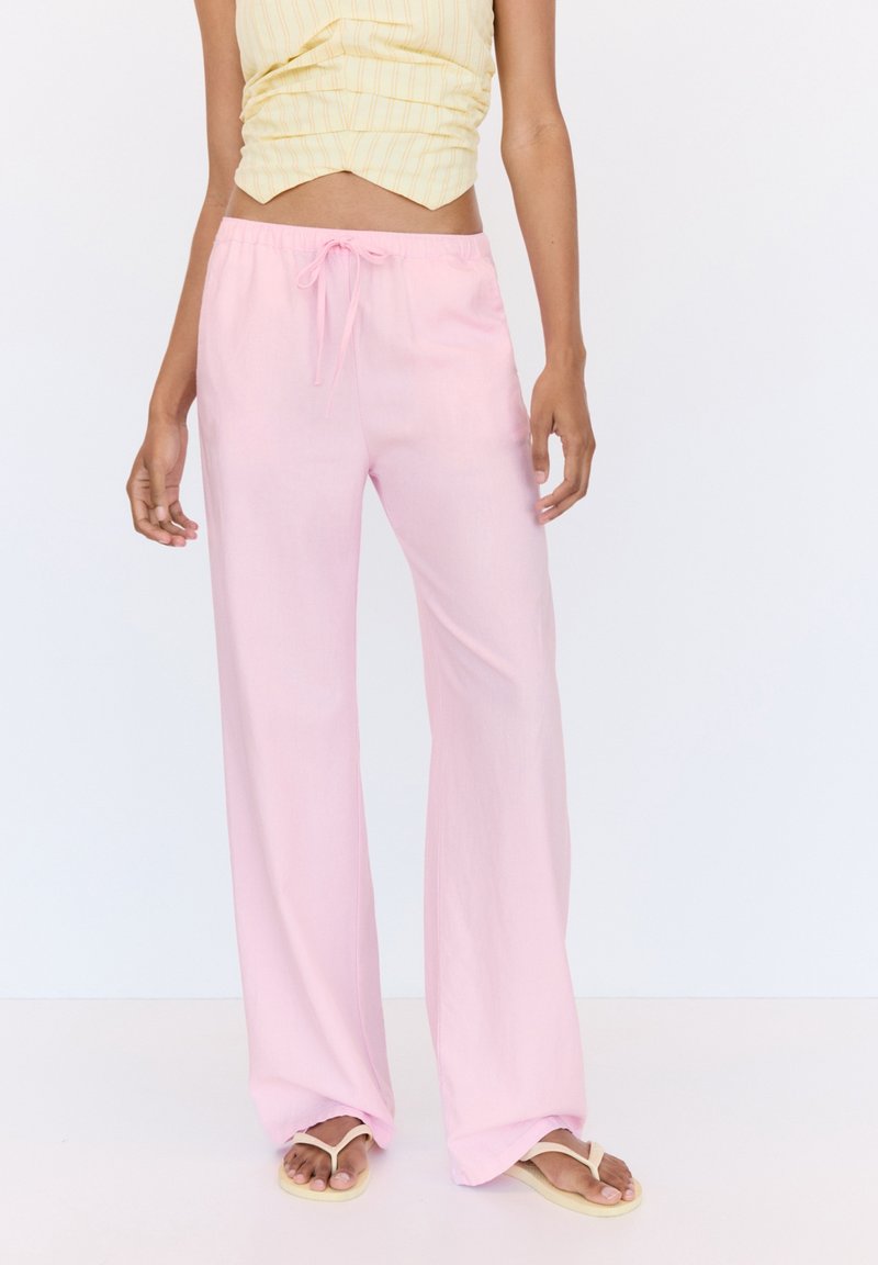 Personne portant un pantalon large rose clair avec cordon, un haut sans manches à rayures jaunes et des tongs beige sur un fond blanc.