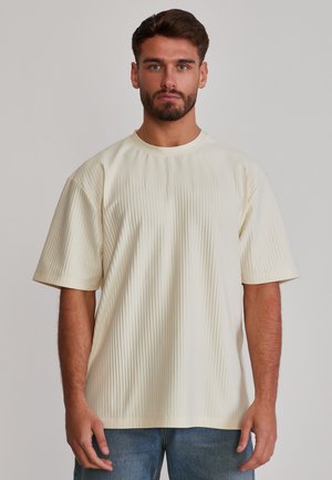 Mann mit braunen Haaren und Bart, der ein cremefarbenes, geripptes Kurzarmshirt und blaue Jeans trägt, vor einem schlichten hellen Hintergrund stehend.