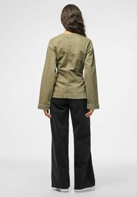 Donna con lunghi capelli mossi, che indossa una giacca verde oliva e pantaloni neri a gamba larga, in piedi di spalle su uno sfondo bianco semplice.