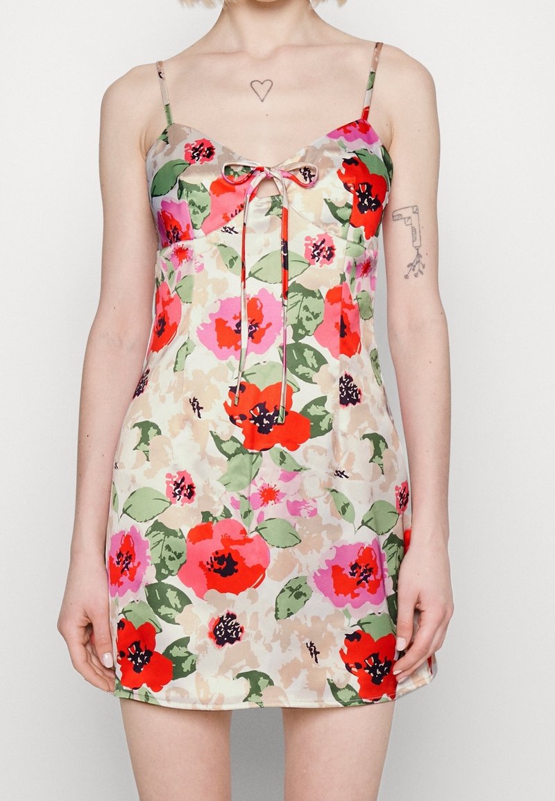 Robe mini florale avec bretelles spaghetti ajustables. Présente un motif de fleurs multicolores sur un fond beige clair, avec un détail de nœud à l'avant.