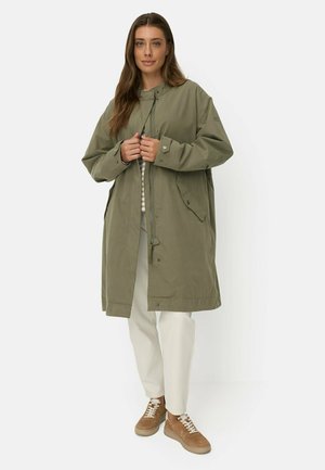 Kvinde iført en oversized olivengrøn frakke, hvide bukser og brune sneakers, stående og lukker frakken foran.
