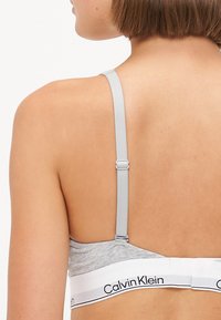 Grijze katoenen bralette met verstelbare grijze banden en een witte elastische band met zwarte "Calvin Klein" branding op de onderband.