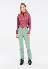 Prilagođena ružičasta majica dugih rukava s crnim točkicama, polu-zip izrez; svijetlozeleni hlače s dva prednja džepa i crnim detaljima; crne skijaške čizme.