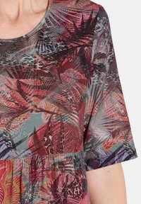 Frau, die ein eng sitzendes, kurzärmliges Oberteil mit einem mehrfarbigen, abstrakten Blumen- und geometrischen Muster in Rot-, Lila- und Grautönen trägt.