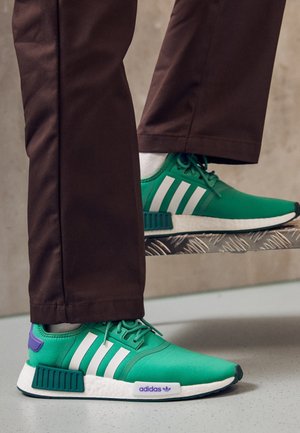 Zielone buty sportowe Adidas z białymi paskami, fioletowymi akcentami, noszone z brązowymi spodniami, jedna stopa na metalowym stopniu, widoczne białe skarpetki.