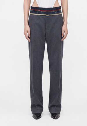 Pantalon gris à jambes larges avec une ceinture multicolore visible, porté avec des chaussures noires pointues et un haut blanc.