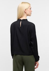 Maglietta nera a maniche lunghe con colletto e polsini arricciati, caratterizzata da un dettaglio a forma di chiave sulla parte posteriore e una texture di tessuto liscia. Indossata con pantaloni verdi.