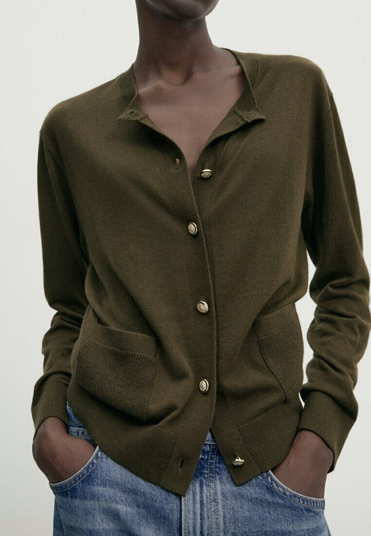 Personne portant un cardigan boutonné vert olive avec des boutons dorés et deux poches avant, associé à un jean bleu et les mains dans les poches.
