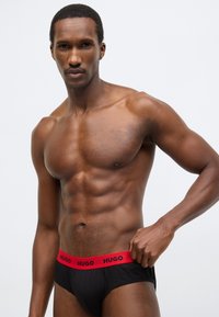 Čierne bavlnené slipy s červeným elastickým pásom s logom "HUGO"; model ukazuje svalnatý horný trup a neutrálny pozadie.