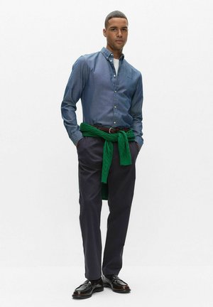 Blå jeansskjorta med knappar, mörka byxor och en grön tröja knuten runt midjan; svarta finskor kompletterar outfiten.