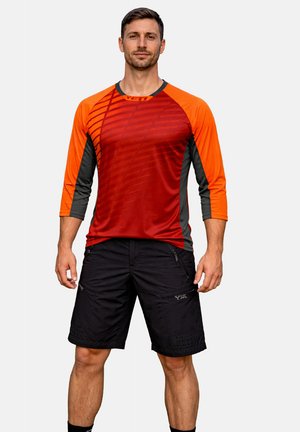 Mann steht vor einem einfarbigen Hintergrund und trägt ein rot-oranges langärmeliges Sportshirt sowie schwarze Shorts bis zum Knie.