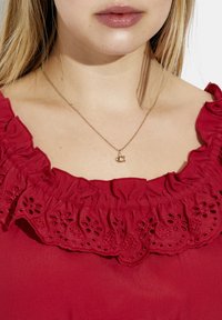 Rød bluse med en flæset halsudskæring og blomster-øjenlåg detaljer. En delikat guldhalskæde med en lille bogstav "C" vedhæng er synlig.