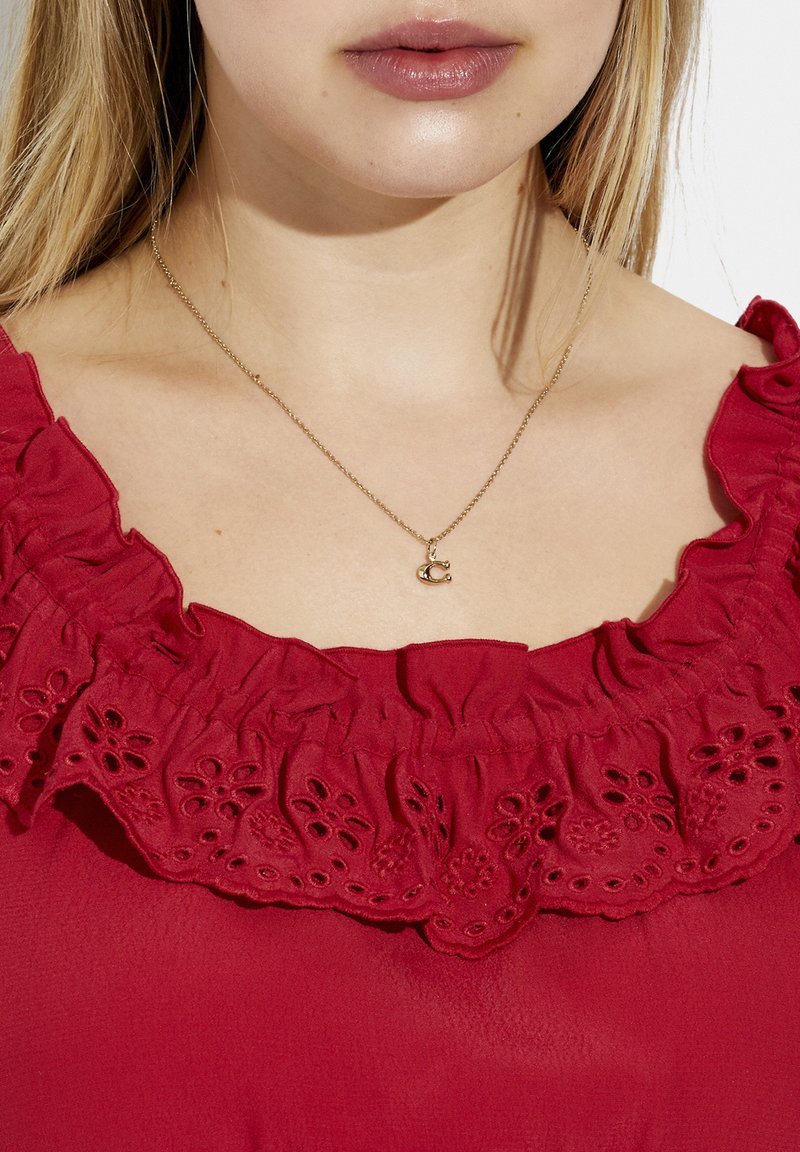 Rød bluse med en flæset halsudskæring og blomster-øjenlåg detaljer. En delikat guldhalskæde med en lille bogstav "C" vedhæng er synlig.