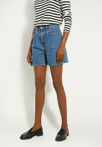 Denim shorts in medium blue with a high waist, featuring a front button closure, five pockets, and visible stitching. Paired with black loafers.