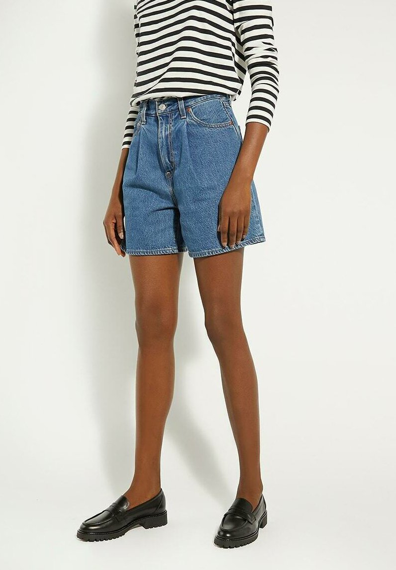 Denim shorts in medium blue with a high waist, featuring a front button closure, five pockets, and visible stitching. Paired with black loafers.
