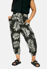 Pantalons noirs avec un motif de feuilles de palmier blanches, une taille élastique, des poignets fuselés et des poches latérales. Portés avec des sandales noires.