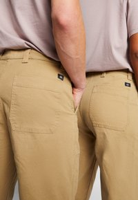 Pantalones caqui con un tejido texturizado, con bolsillos traseros que tienen etiquetas negras en contraste, vistos desde la parte de atrás. Diseño sencillo y de corte recto.