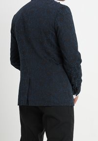 Uomo che indossa un blazer a motivi blu scuro e pantaloni neri, in piedi con la schiena rivolta alla fotocamera su uno sfondo bianco.